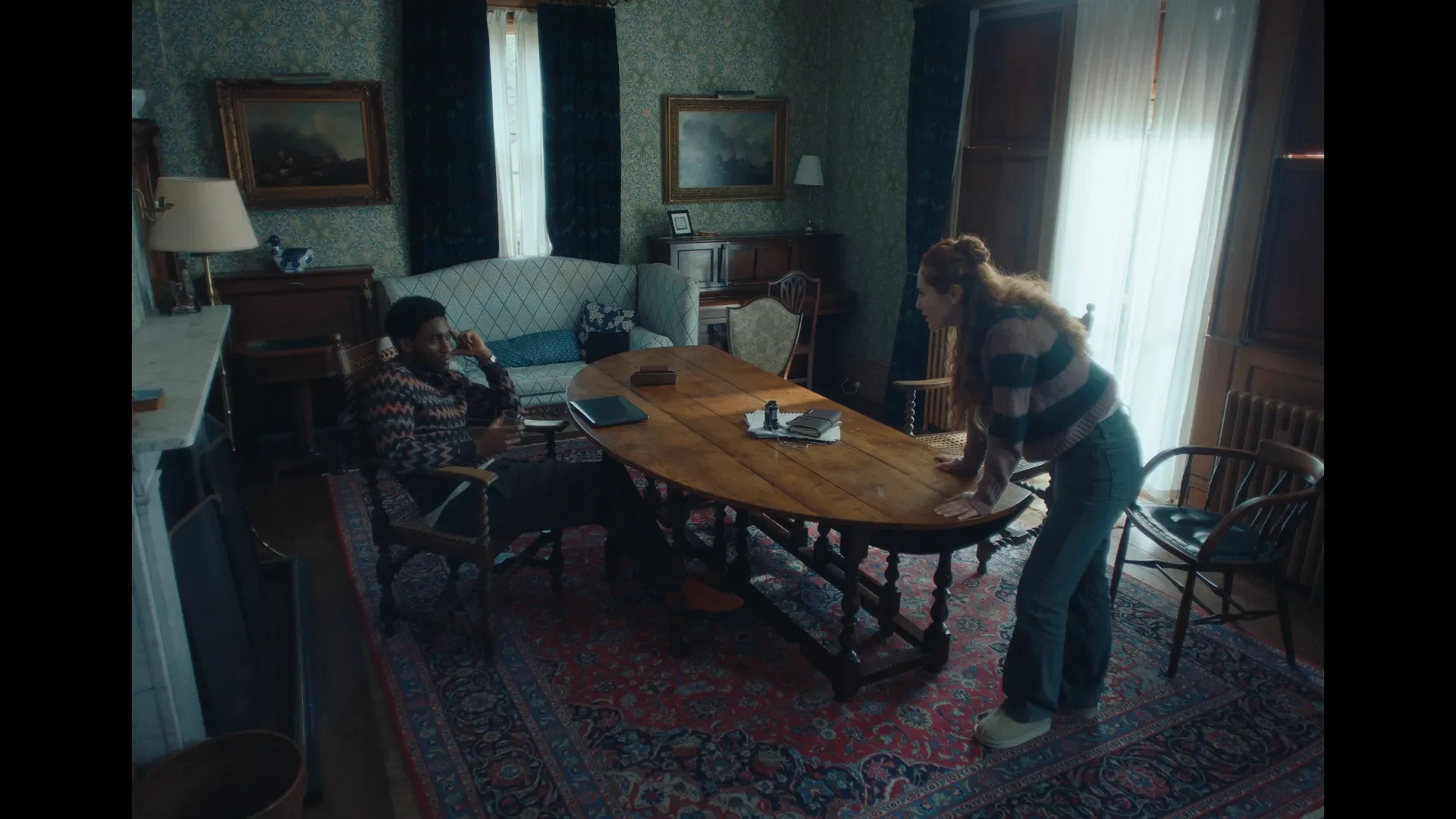 The image shows two people, a man sat down at a table and a woman leaning on the table looking straight at him.