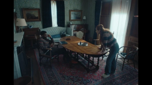 The image shows two people, a man sat down at a table and a woman leaning on the table looking straight at him.