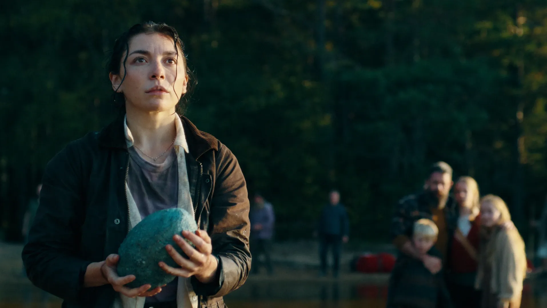 A woman stands holding a large blue egg. A family group huddled together are standing behind her