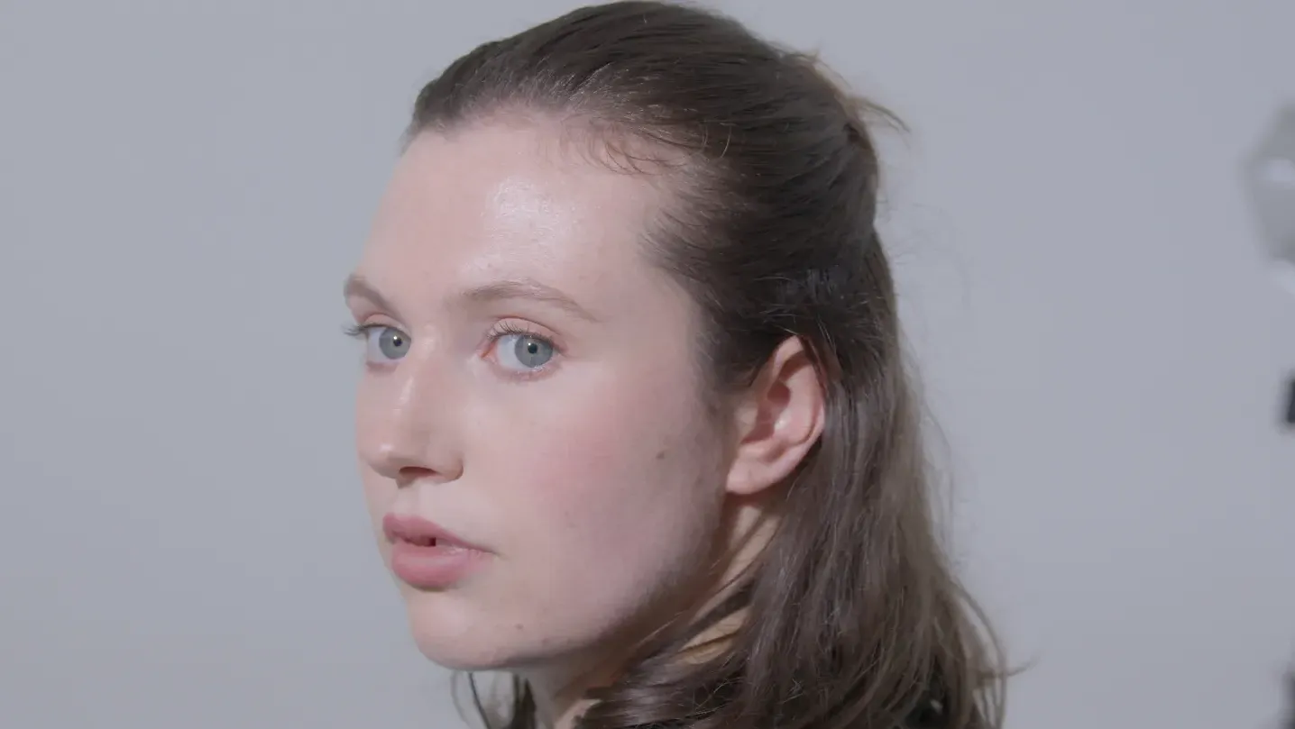 A woman with her head turned around looking behind her. She is in a plain white studio background, she has medium length brown hair half tied up half let down, and has blue eyes