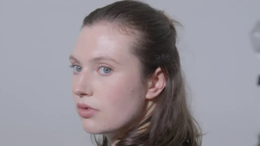 A woman with her head turned around looking behind her. She is in a plain white studio background, she has medium length brown hair half tied up half let down, and has blue eyes