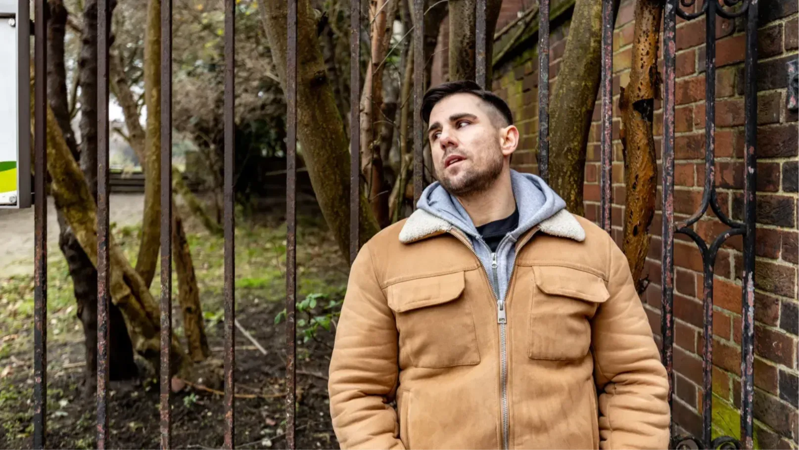 A man wearing a light brown jacket and a grey hoodie rests against a park railing.