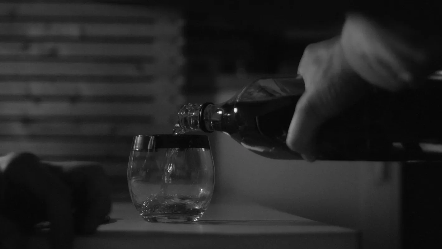 A close up shot of a hand pouring whisky into a glass on a kitchen table.