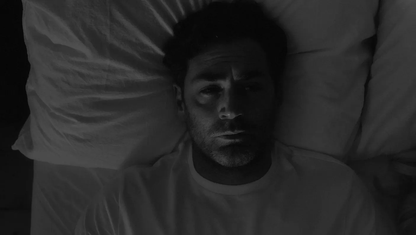 A man lying in bed at night, looking up at the ceiling, next to his wife.