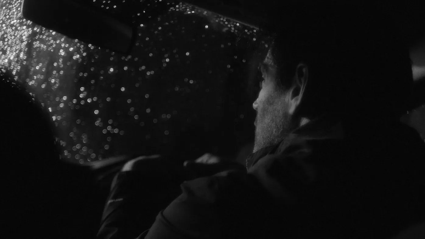 A man sitting inside his car at night, with the windows covered in raindrops.