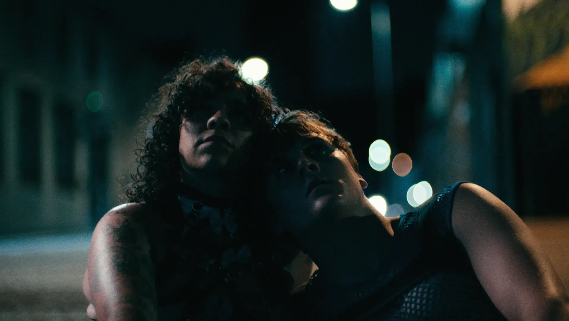 Two people sat in the street at night time. They are dressed in club-wear. Shadows partially cover their faces and lights blur in the background.