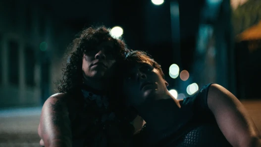 Two people sat in the street at night time. They are dressed in club-wear. Shadows partially cover their faces and lights blur in the background.