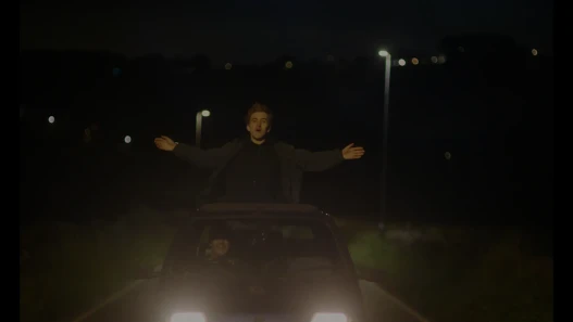 A young man is standing out of car sun-roof as another young man drives the car