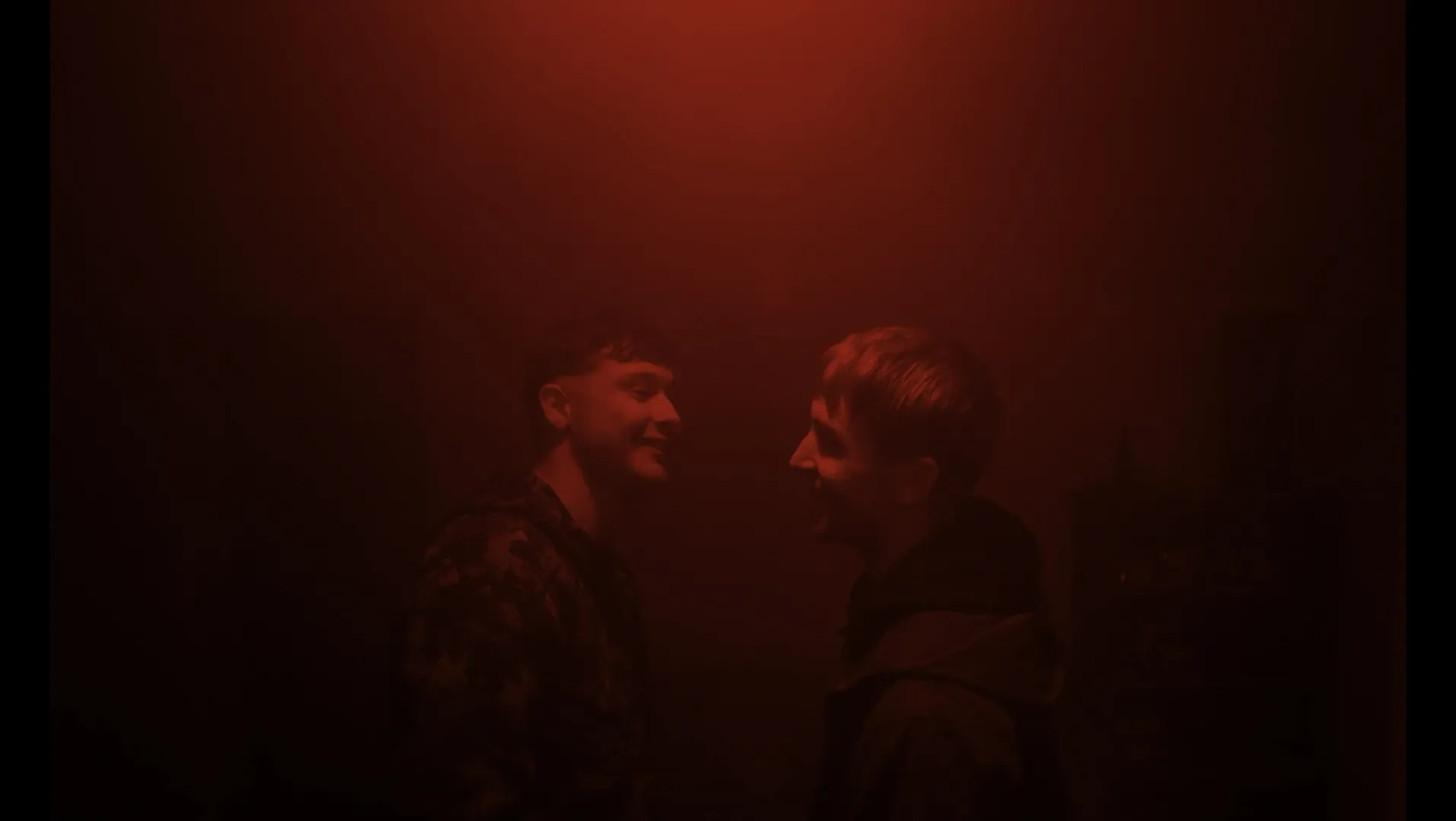 Two smiling young men stand in red hazy room