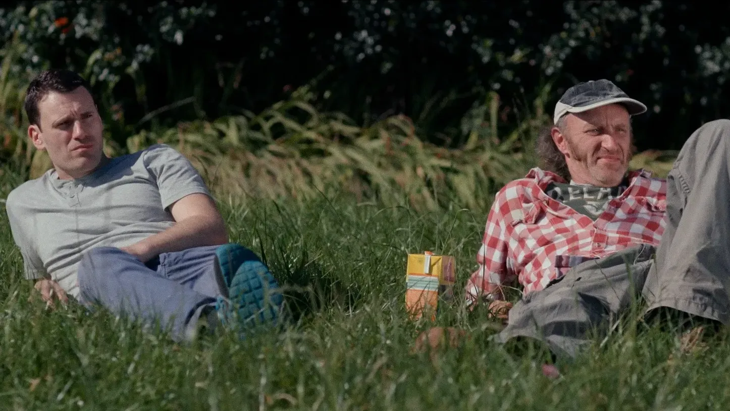 Two men reclining in a field. There is a carton of juice between them.