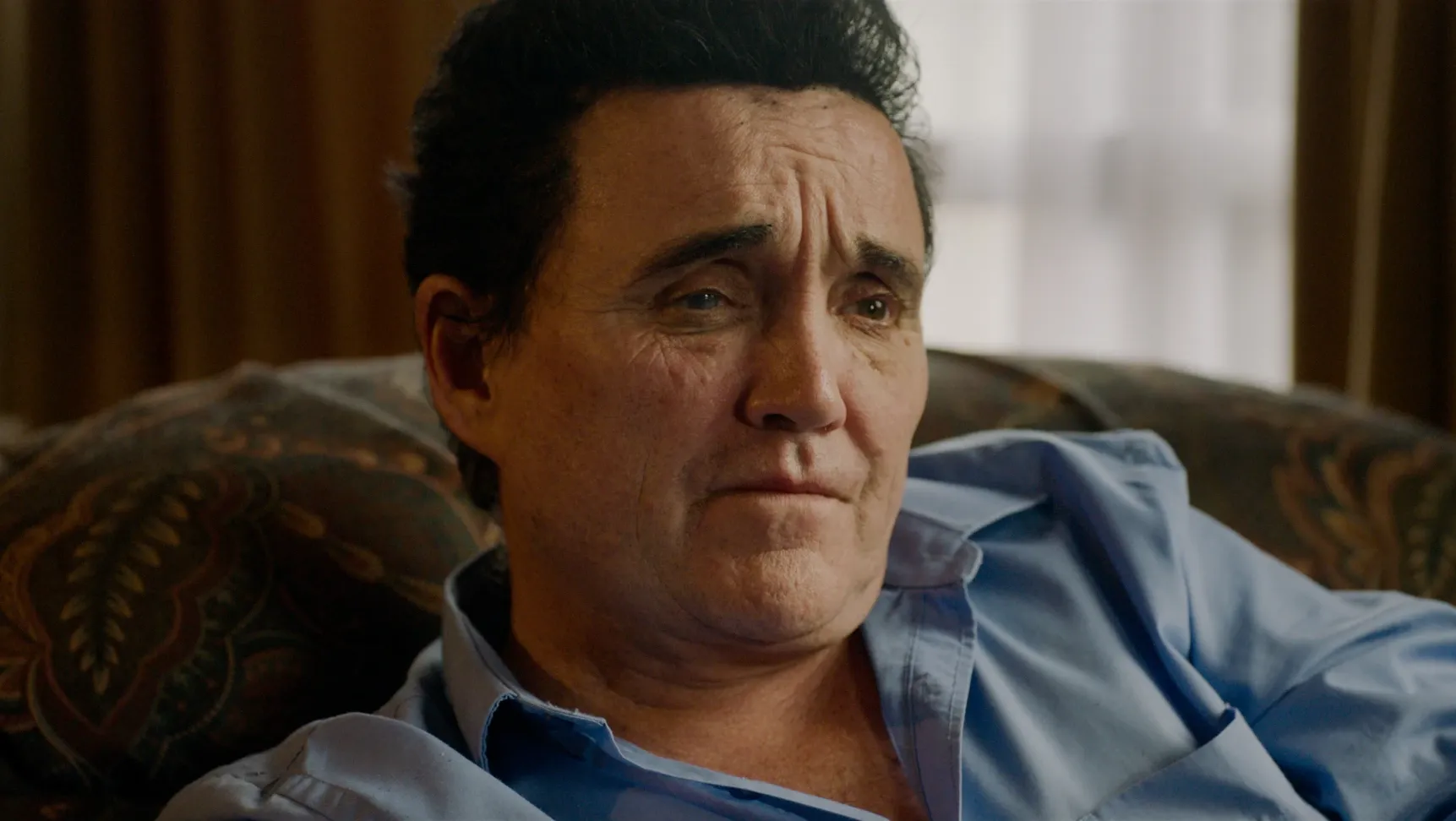 A close up shot of a man sat on a sofa wearing a blue shirt.