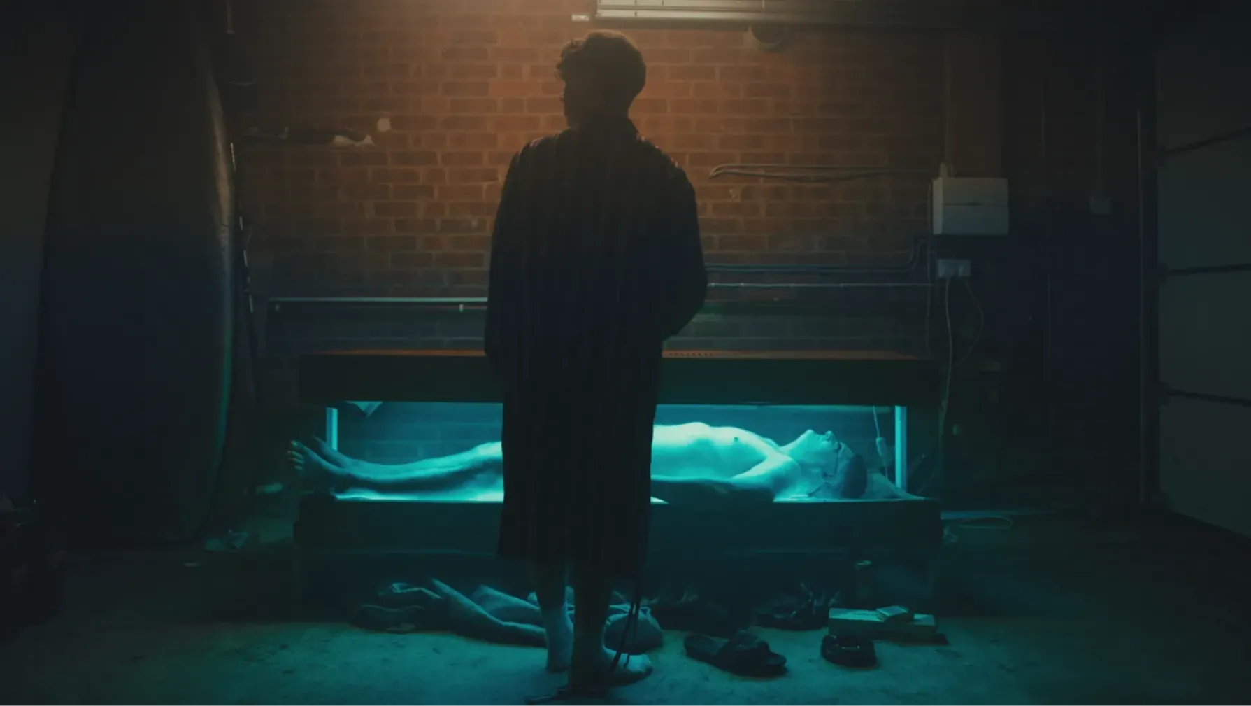 Someone lies on a sunbed, shrouded in blue light in a garage, whilst someone else looks on