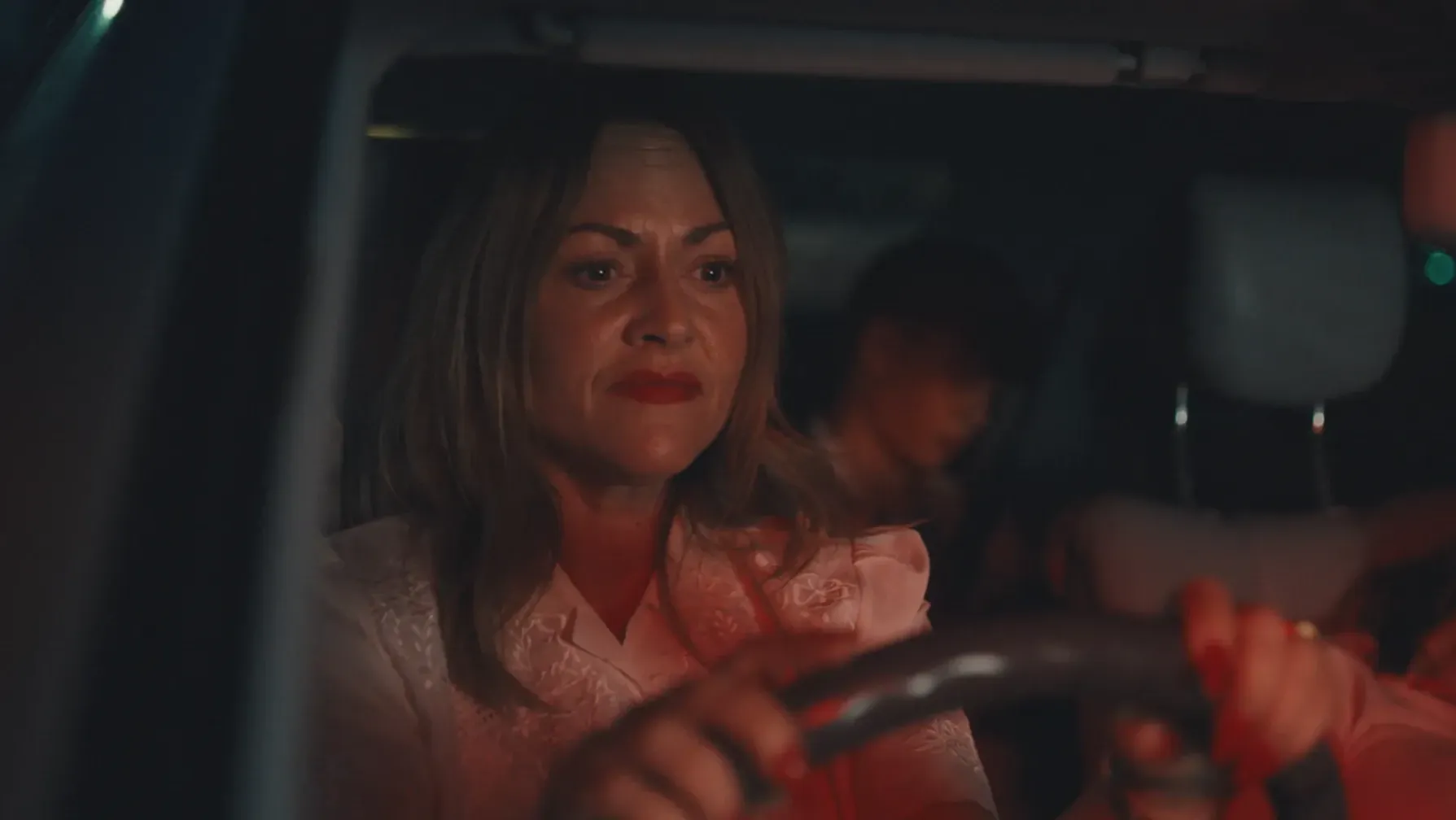 A mother driving and looking tense, her teenage son in the back of the car half in shadow.. 