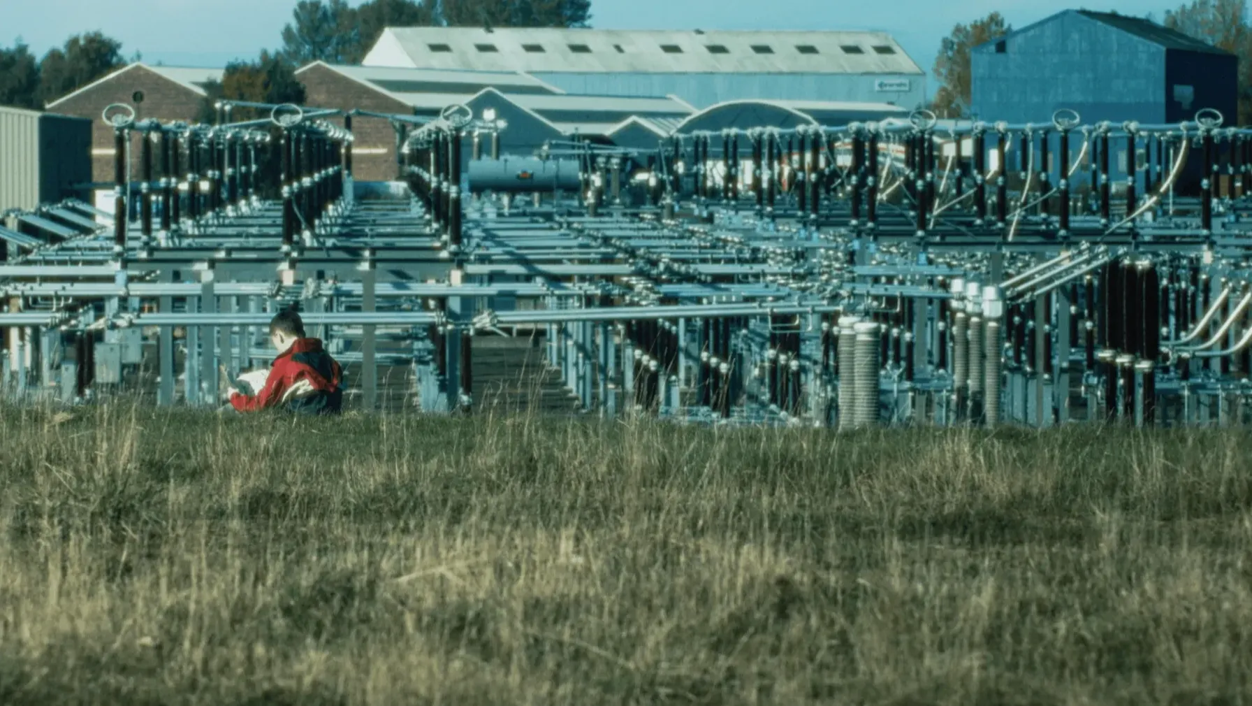 A wide shot image of a boy sitting in a field with his back to the camera, reading a magazine. In the distance behind him is a tangle of pylons and industrial buildings