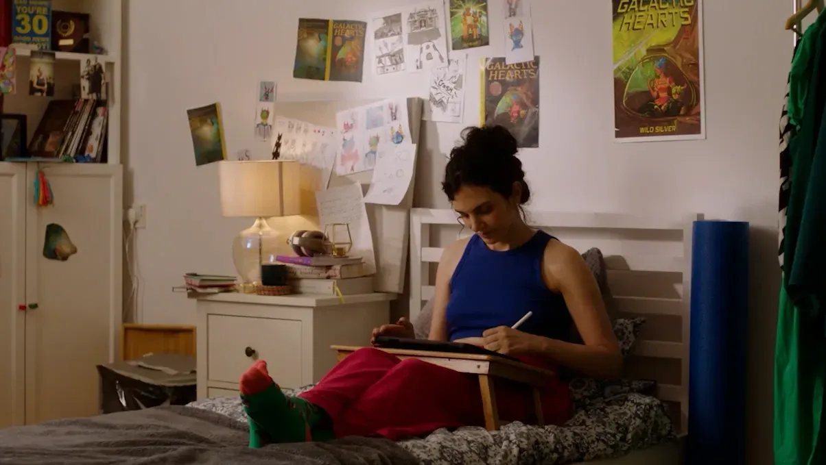 British-Mexican woman sat on her bed with a tray on her drawing a comic.