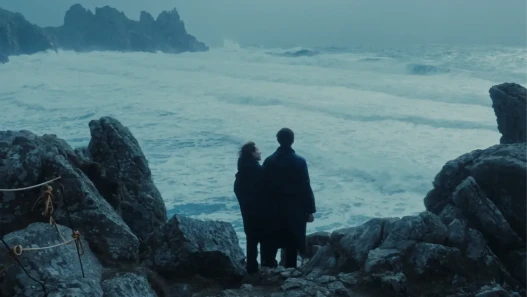 A woman and a man stand on a stormy cliff edge looking out to sea.