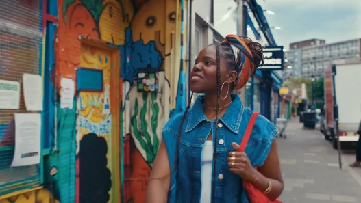 A woman walking down an East London street, wearing a denim jacket, red handbag and hooped earrings.