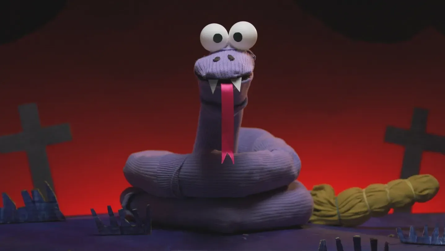 A sock puppet snake stares into the camera in a cardboard graveyard.