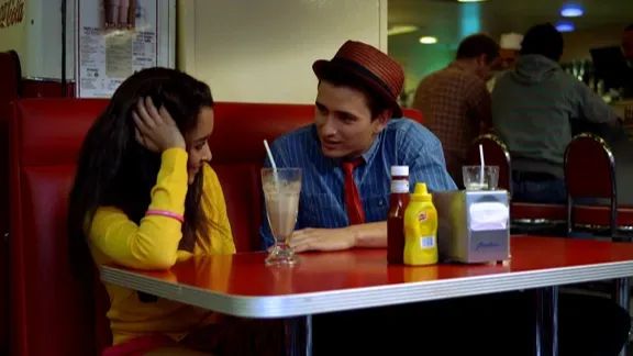 A young boy wearing a trilby hat, and girl sit in a colourful diner with a milkshake on the table, engaged in a flirtatious interaction.