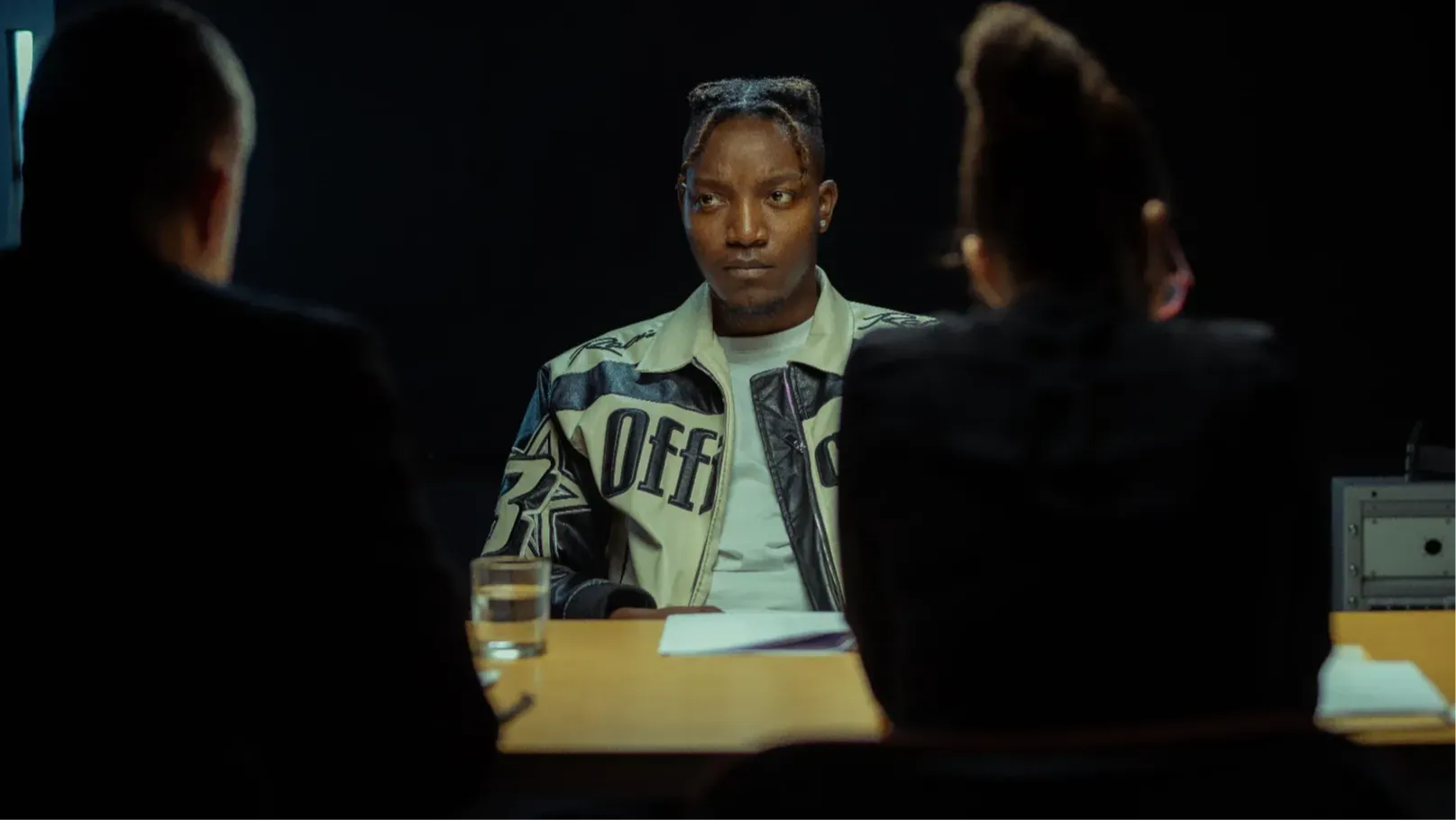 A young black man faces two detectives, who are viewed from behind.