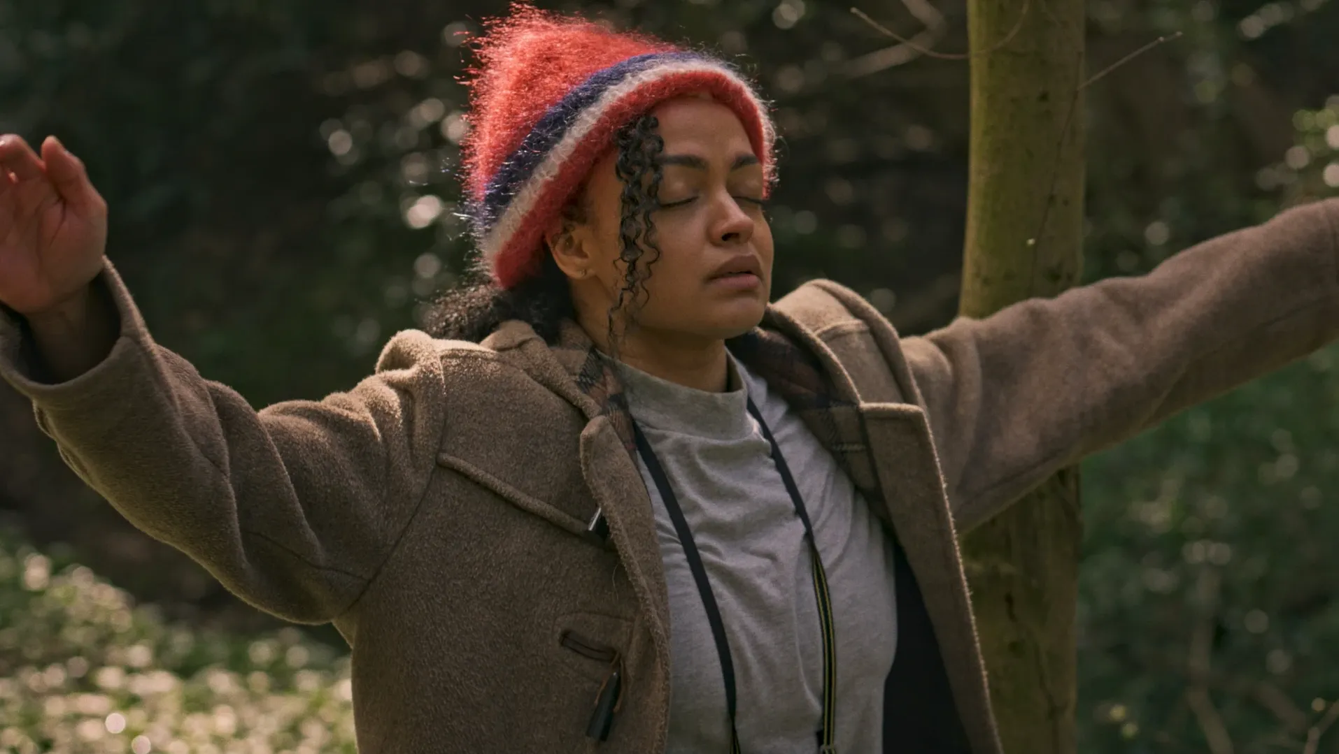 A mixed race woman in a red hat and brown coat raises arms with eyes closed