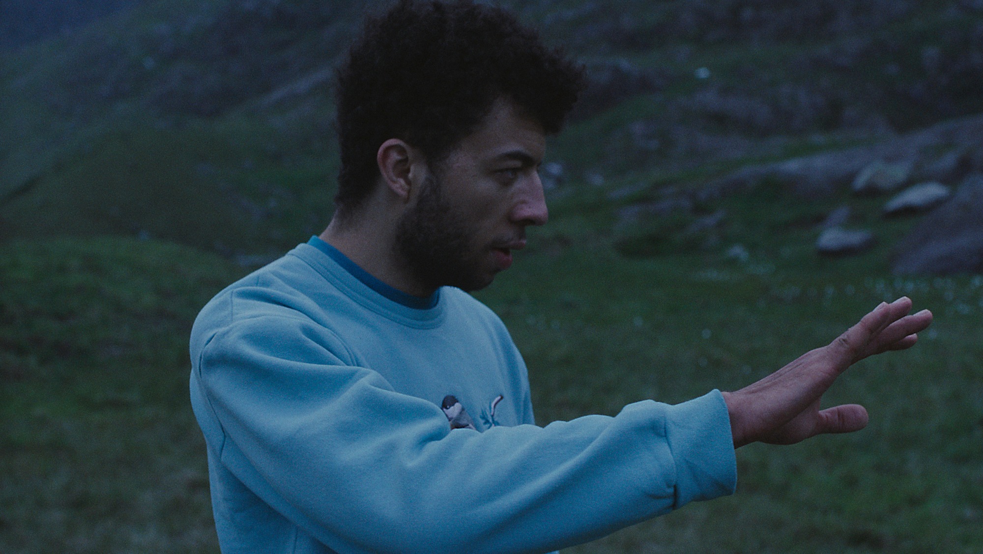 Outside at dusk, a young mixed-raced man lifts his hand.