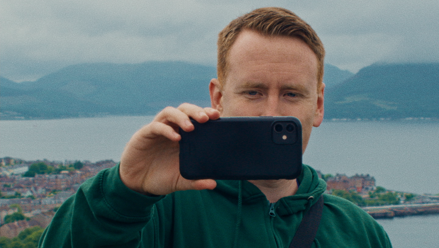 Outdoors. A man holds a mobile phone in front of him and records using the phone's camera.