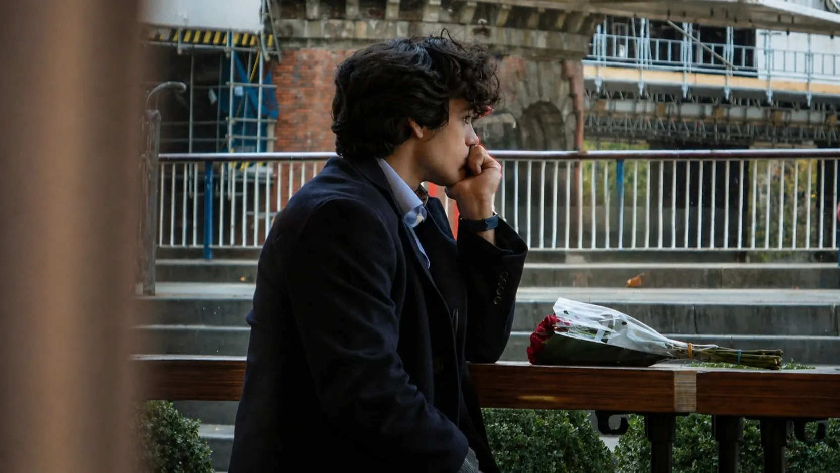 A young man, sits alone in a cafe, deep in thought. A bouquet of disregarded roses in front of him.