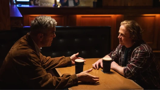 Two men face each other across a table with a pint in a pub.