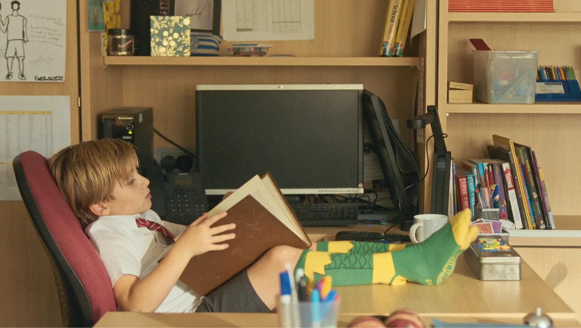 A young boy seated in an office chair with his feet on the desk is reading a book.