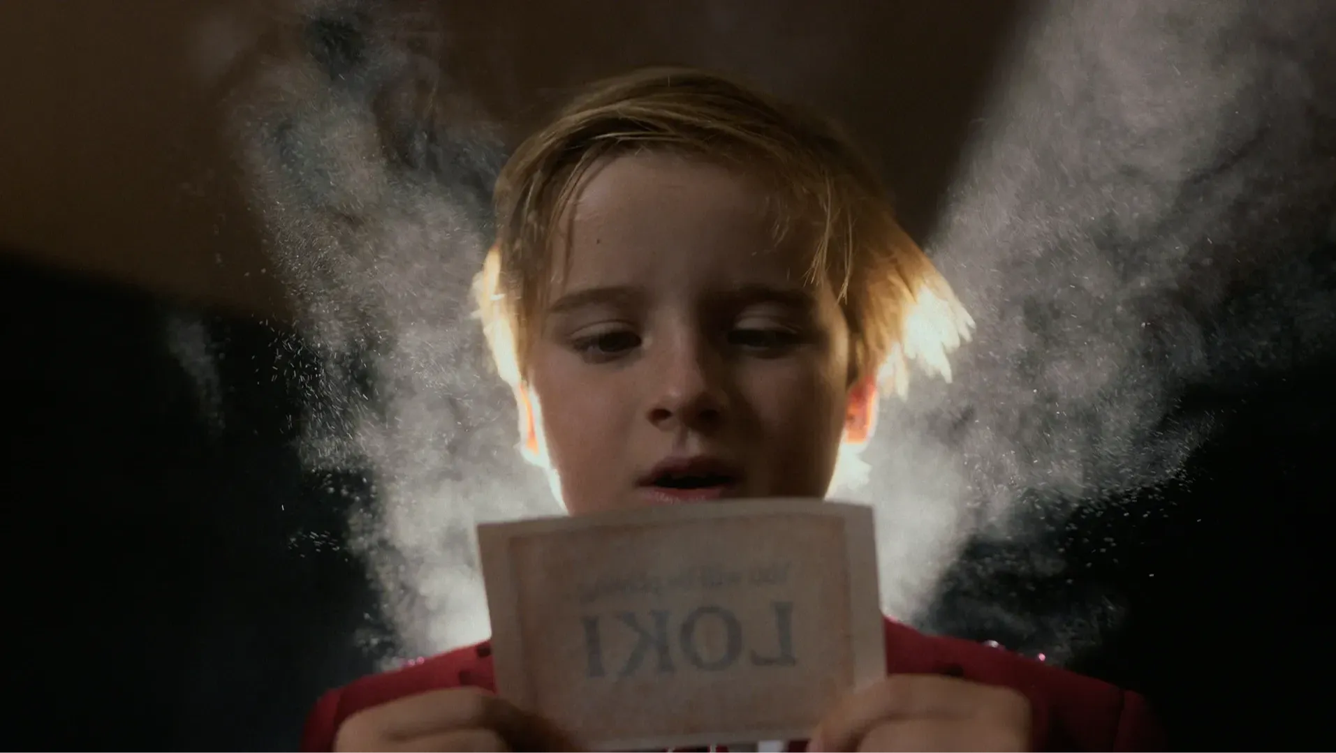 A young boy with light shining behind him reading a piece of paper with the word "Loki" on it