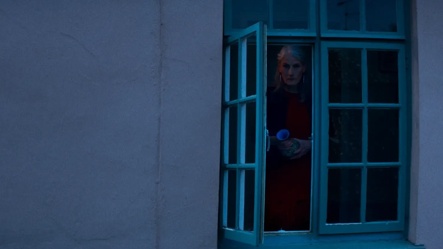 At dusk a woman stands at window holding a vase