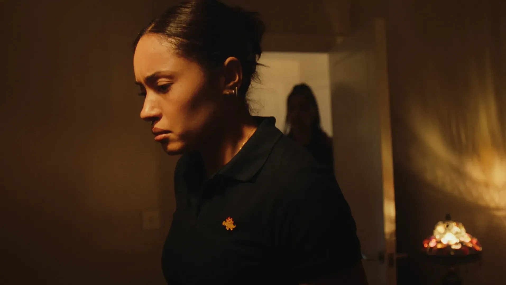 A woman standing the foreground with a furrowed brow, another woman is standing silhouetted in the background doorway.