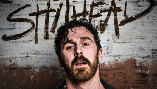 A man looks straight into the camera. He is standing in front of a wall with graffiti on it.