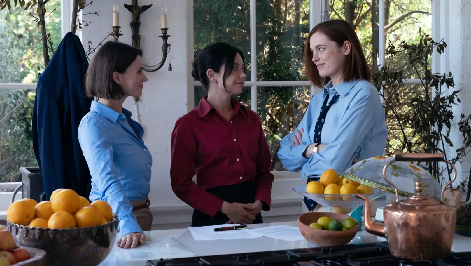 Three women wearing shirts are standing in a kitchen with windows behind them.