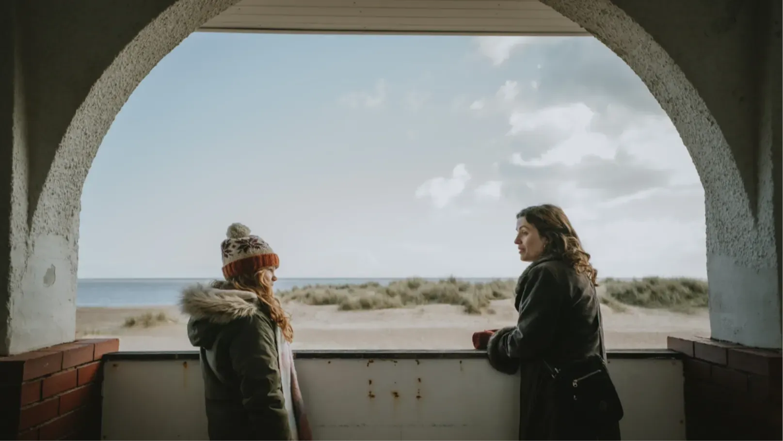 A girl and a woman talk with a view of a beach and the sea behind them.