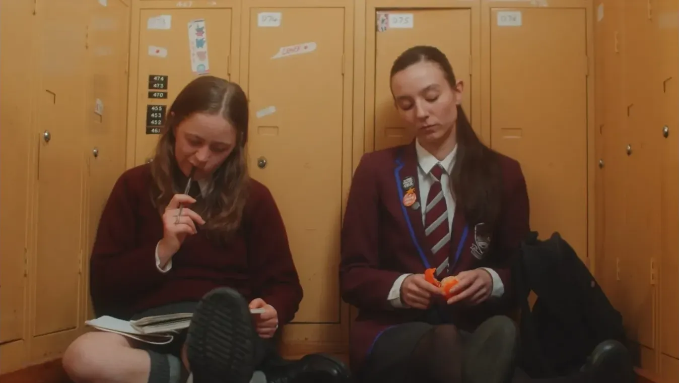 Two girls in maroon school uniform sit against mustard yellow lockers doing homework and sharing an orange.