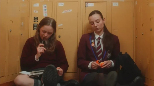 Two girls in maroon school uniform sit against mustard yellow lockers doing homework and sharing an orange.