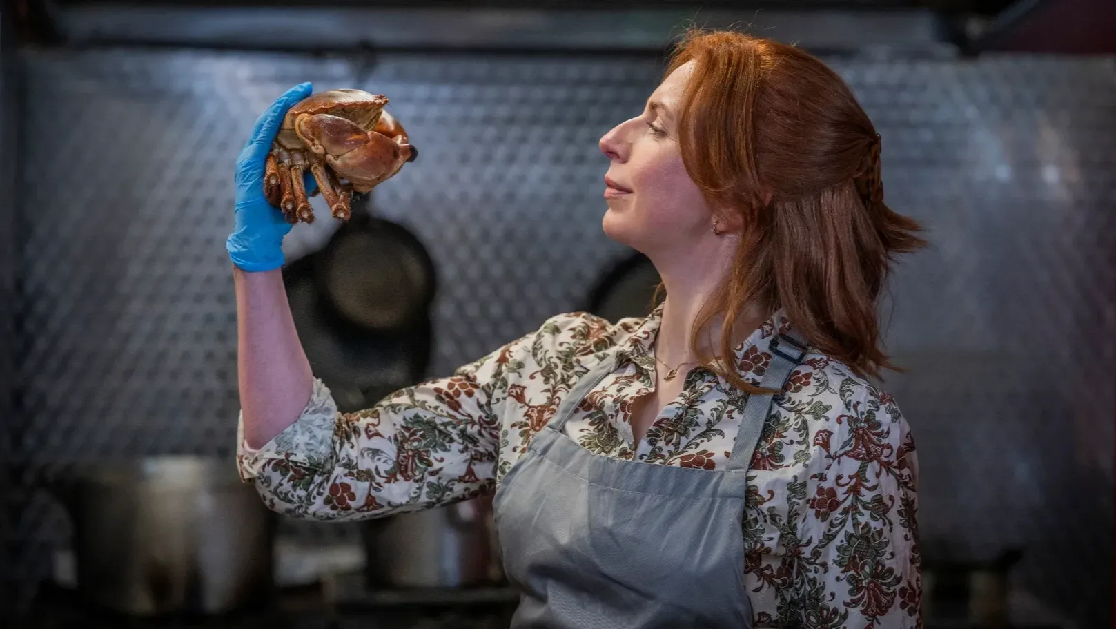 A woman wearing and apron and protective gloves in a kitchen is holding a crab