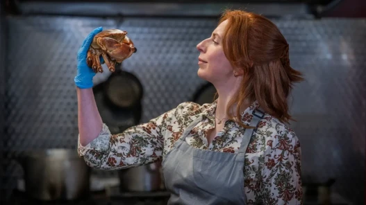 A woman wearing and apron and protective gloves in a kitchen is holding a crab