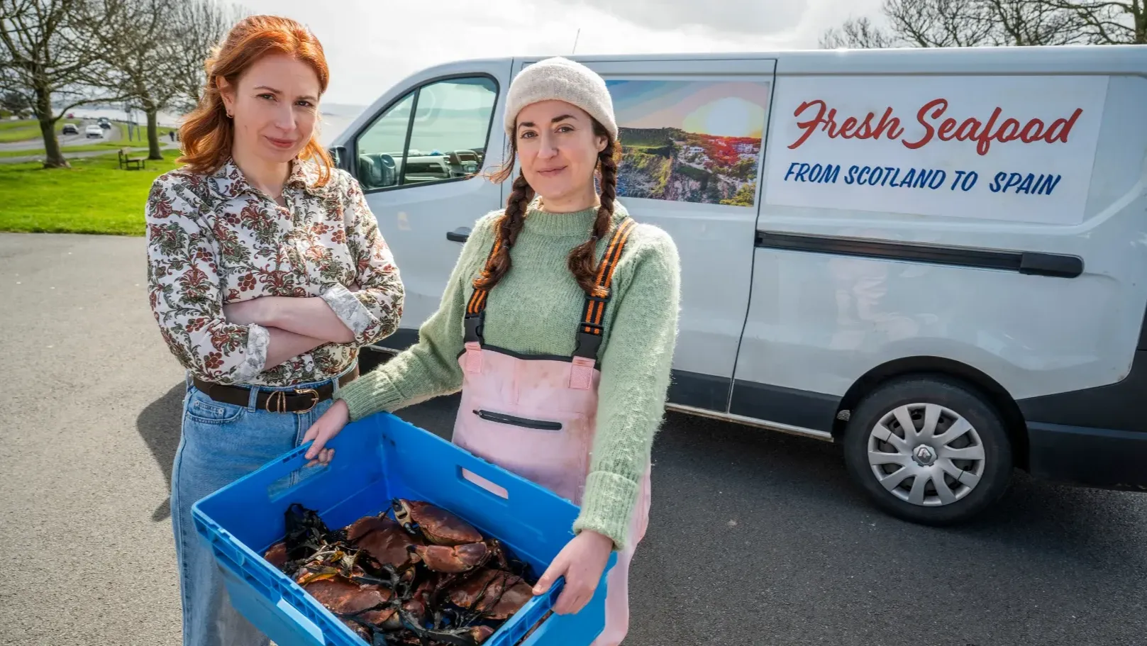 Two women are standing outside in front of a branded van "Fresh Seafood. From Scotland to Spain". One of the women ins holding a crate of crabs.
