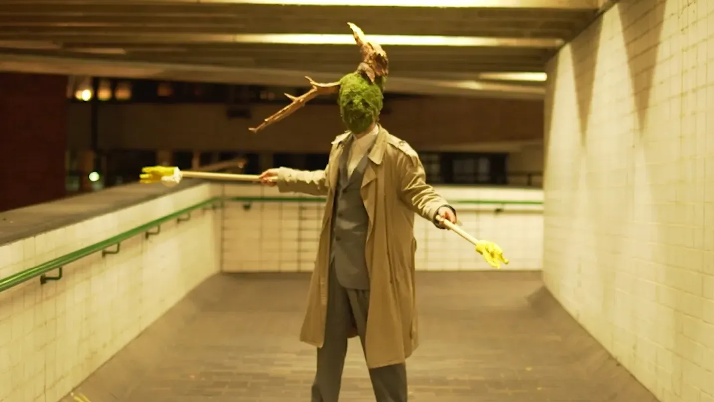A man-like figure with a moss covered face and wooden branches stick out of their head stood in a suit and mackintosh overcoat holding poles with yellow hands on the end.