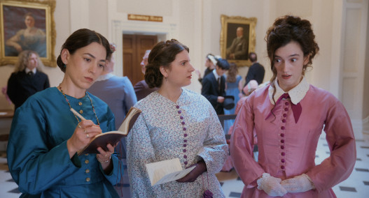 Three women stand together. One woman in blue writing in a notebook, another woman in white and blue patterned dress holding a booklet and a third lady in a pink dress looking surprised. 