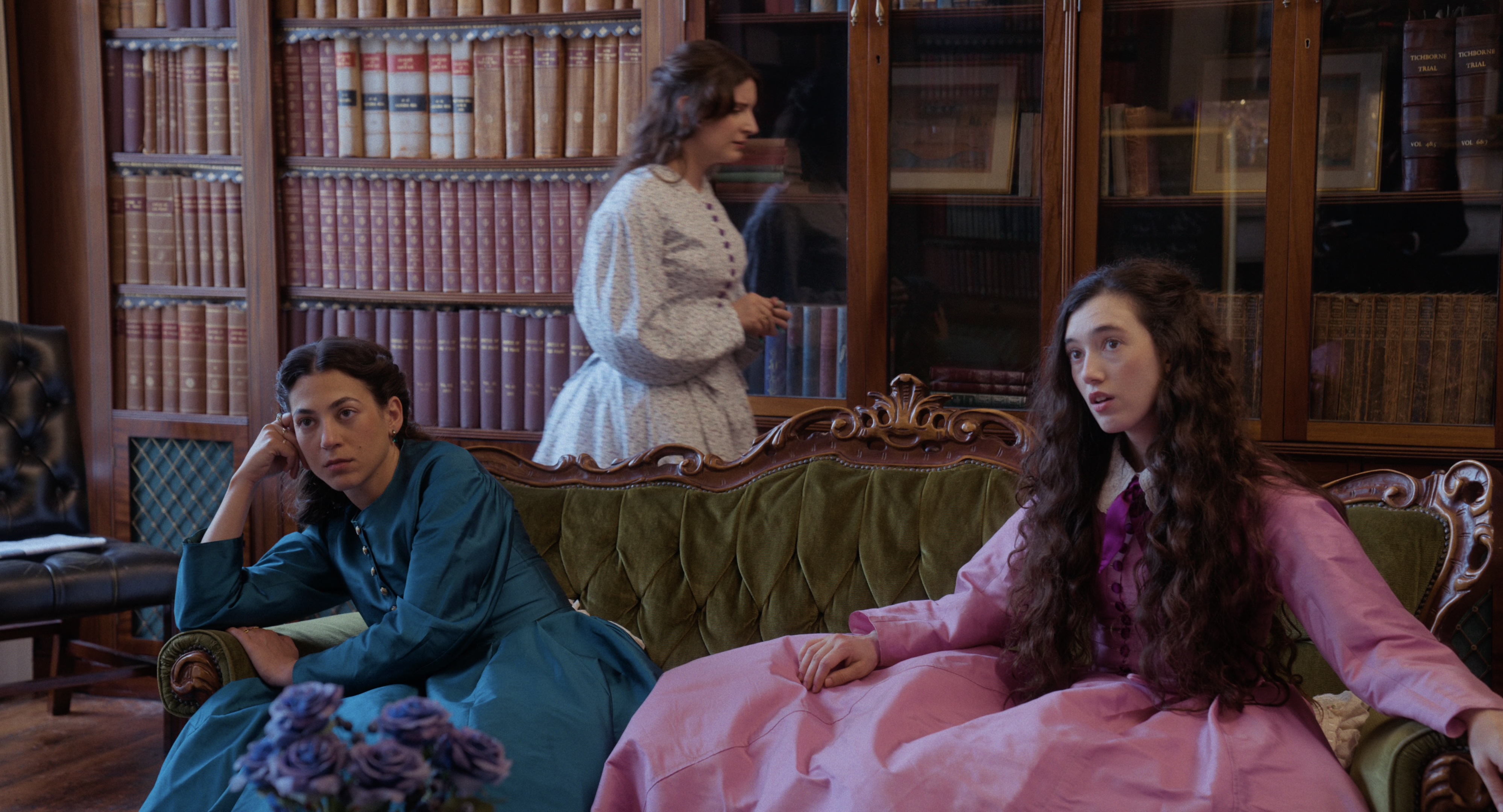 Two women, left in a blue dress and right in a pink dress sit on a green sofa with a woman in white walking behind. 