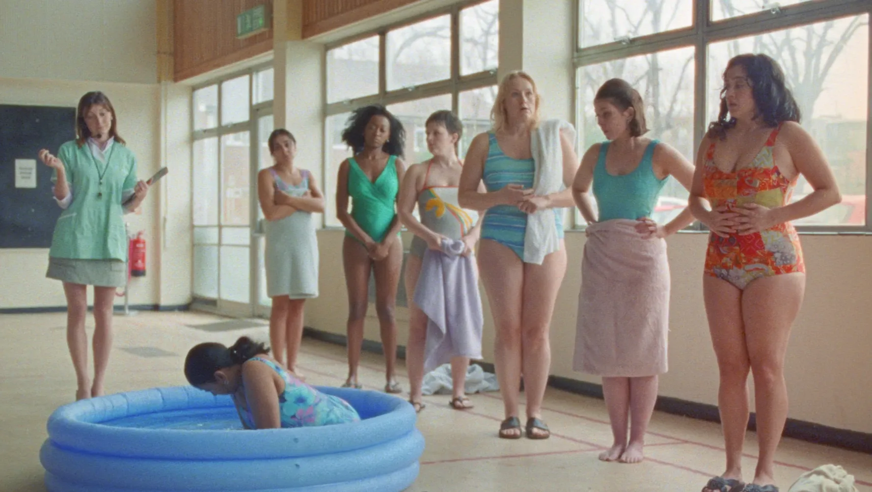 A woman sits in a paddling pool, while a line of women wait on the sidelines
