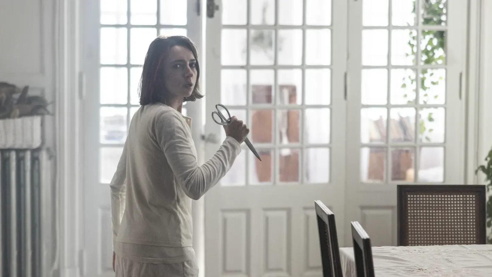 A woman with dark brown hair holds a pair of scissors and looks frightened in a big house.