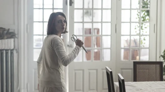 A woman with dark brown hair holds a pair of scissors and looks frightened in a big house.