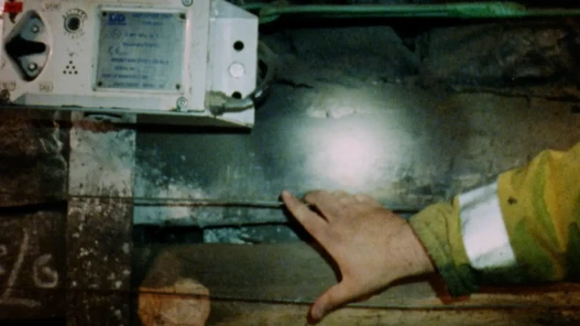 A hand rests on two metal wires in an underground mine. An electrical box is above. A head torch shines onto the rock face.