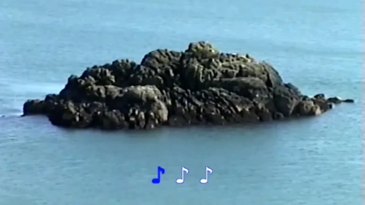 A rocky outcrop in the ocean, karaoke-style music notes in lower half of frame