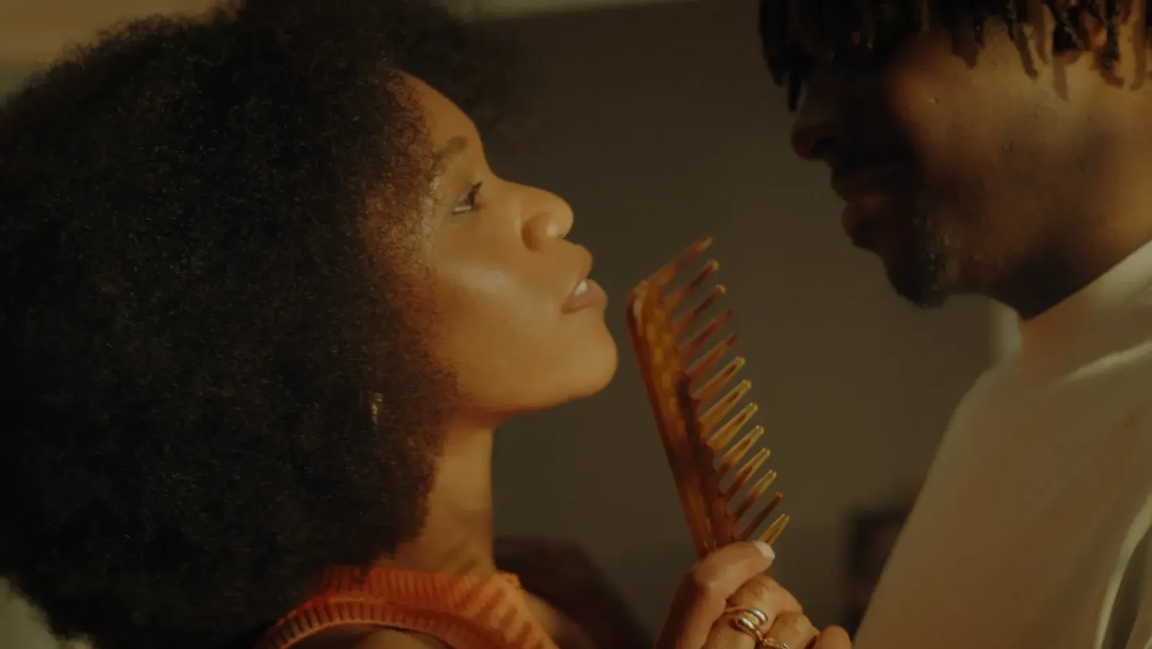A Black woman wearing her hair in an afro holds a Wide Tooth Comb stood in front of a Black man in a white T-shirt who wears his hair in short locs.